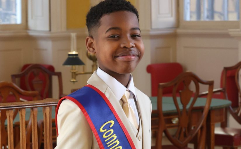 CT's 2025 kid governor Keudy Martinez wearing a off-white suite with a blue and red sash that says "Connecticut".